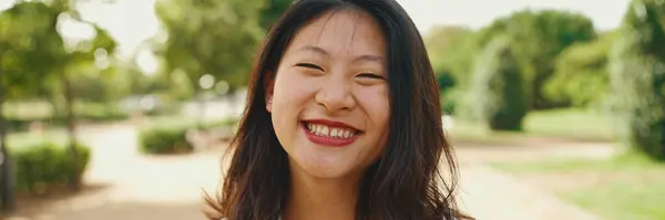 Close-up young Asian smiling woman with long brown hair wearing white t-shirt posing for the camera in the park, Panorama. The girl opens her eyes and smiles at the camera