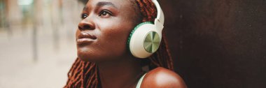 Kulaklıklı, Afrika örgülü, sokakta müzik dinleyen güzel bir kadının yakın çekimi, Panorama. Şık kız gözleri kapalı müzik dinliyor.