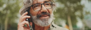 Panorama, orta yaşlı, dost canlısı, gri saçlı ve sakallı bir adam cep telefonunu kullanarak günlük giysiler giyiyor. Gözlüklü, dışarıda cep telefonuyla konuşan olgun bir beyefendi.
