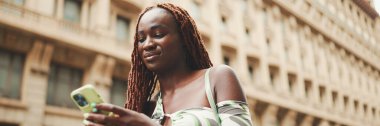 Üzerinde Afrika örgüleri olan gülümseyen muhteşem bir kadın yolun yanındaki kaldırımda duruyor ve cep telefonu kullanıyor, Panorama. Şık kız akıllı telefondan mesaj yazıyor.