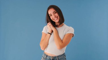 Beyaz bluz giymiş, gülümseyen genç bir kadın. Avuçlarını katlayan kameraya bakıyor. Stüdyoda mavi arka planda çok yalnız.