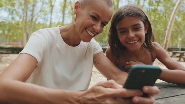 Parkta cep telefonuyla oturan mutlu anne ve kız.