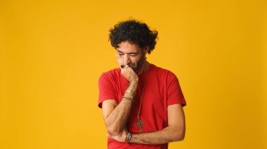 Kıvırcık saçlı, kırmızı tişörtlü hoşnutsuz bir adam. Stüdyoda sarı arka planda izole edilmiş bir kameraya bakarken anlaşmazlık gösteriyor.