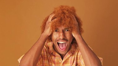 Yakından, şaşkın stil kıvırcık saçlı adam, kameraya büyük gözlerle bakıyor, diyor ki vay canına, ellerini başının üstüne kaldırıyor, stüdyonun kahverengi arka planında izole edilmiş.