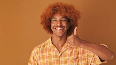 Yaklaş, şık kıvırcık çocuk, kameraya bakıyor, başparmak hareketi gösteriyor, stüdyoda kahverengi arka planda izole edilmiş.