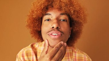 Tarz sahibi kıvırcık çocuk, kameraya aynada bakar gibi bakıyor, stüdyonun kahverengi arka planında izole edilmiş.