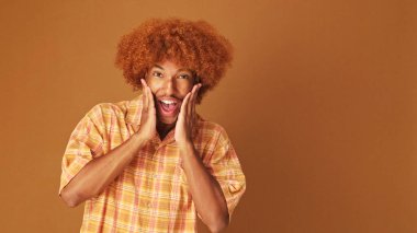 Şaşırmış stil sahibi kıvırcık çocuk kameraya şaşırmış bir şekilde bakıyor. Model mekan, stüdyonun kahverengi arka planında izole edilmiş.