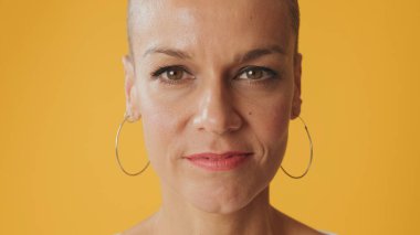 Stüdyoda sarı arka planda izole edilmiş kameraya bakan tüysüz genç bir kadının yakın plan portresi.