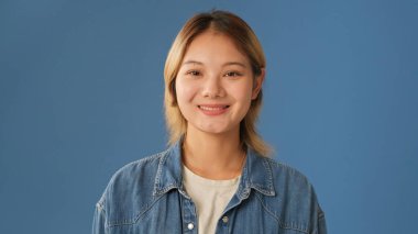 Kot gömlek giymiş, kameraya bakıp gülümseyen, stüdyoda mavi arka planda izole genç bir kadının yakın çekimi.