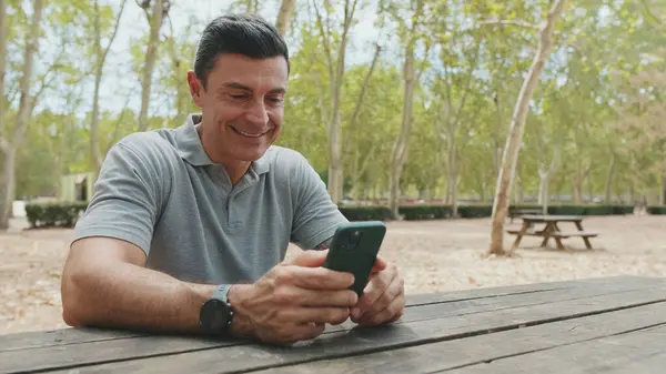 Gri tişört giyen orta yaşlı bir adam park bankında otururken cep telefonu kullanıyor.