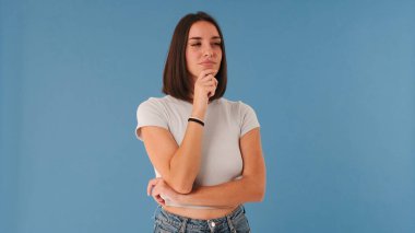 Beyaz bluzlu güzel esmer kadın kameraya bakar. Plan hakkında düşünür. Stüdyoda mavi arka planda izole edilmiş bir fikir.