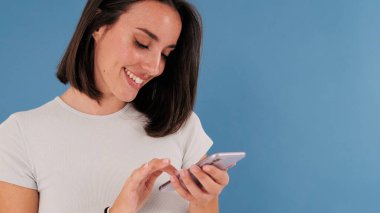 Yaklaş, güzel gülümseyen esmer kadın. Beyaz elbiseli. Stüdyoda mavi arka planda izole edilmiş cep telefonu kullanıyor.