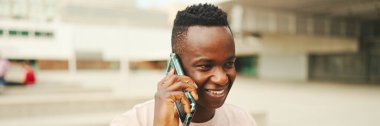 Üniversitenin dışında oturan gülümseyen Afrikalı genç bir öğrenci cep telefonuyla konuşuyor, Panorama.