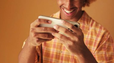 Yakın plan, adamın elleri çevrimiçi oyunlar oynarken cep telefonu tutuyor, kahverengi arka planda izole edilmiş.