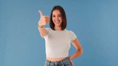 Beyaz giysili güzel esmer kadın kameraya bakar ve stüdyoda mavi arka planda izole bir şekilde başparmağını kaldırır.