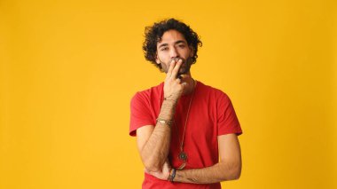Dalgın, kıvırcık saçlı, kırmızı tişörtlü, çenesini ovuşturan ve stüdyoda sarı arka planda yaşayan çekici bir adam.