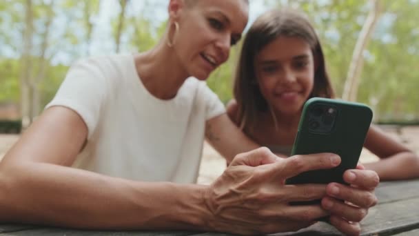 携帯電話を使用して公園に座っている幸せな母親と娘 背景はぼやけ — ストック動画