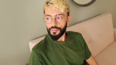 Yakın plan, sakallı genç sarışın adam rahat bir oturma odasında koltukta oturuyor.