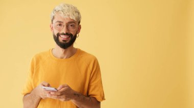 Gözlüklü, sarı tişörtlü, cep telefonuna SMS yazan, sarı arka planda stüdyoda izole edilmiş biri.