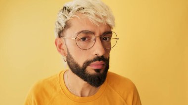 Gözlüklü, sarı tişörtlü, kameraya aynada bakar gibi bakan, stüdyoda sarı arka planda olan bir adamın yakın çekimi.