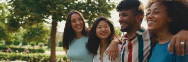 Mutlu çoklu etnik çeşitlilikte genç insanlar yaz günü açık havada kucaklaşarak yürürler, Panorama. Bir grup arkadaş, şehir parkında yürürken neşeyle konuşuyor ve gülüyorlar.