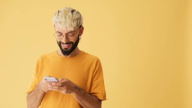 Gözlüklü, sarı tişörtlü, cep telefonuna SMS yazan, sarı arka planda stüdyoda izole edilmiş biri.