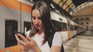 Yaklaş, genç bayan tren istasyonunun platformunda dururken cep telefonunu kaydırıyorsun.