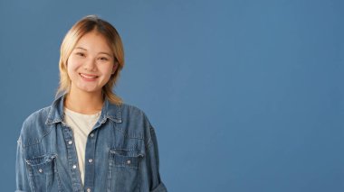 Kot gömlek giymiş genç bir kadın kameraya bakıyor ve gülümsüyor. Stüdyonun mavi arka planında izole edilmiş bir şekilde yer kaplıyor.