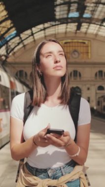 Genç bir kadın elinde cep telefonuyla tren istasyonunun platformu boyunca yürüyor.