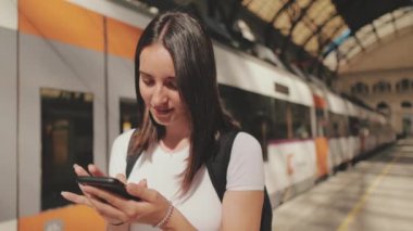 Yaklaş, genç bayan tren istasyonunun platformunda dururken cep telefonunu kaydırıyorsun.