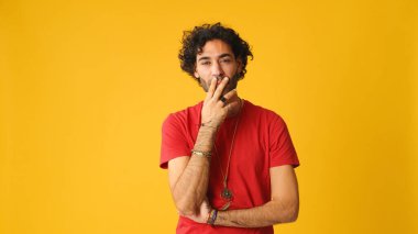 Dalgın, kıvırcık saçlı, kırmızı tişörtlü, çenesini ovuşturan ve stüdyoda sarı arka planda yaşayan çekici bir adam.