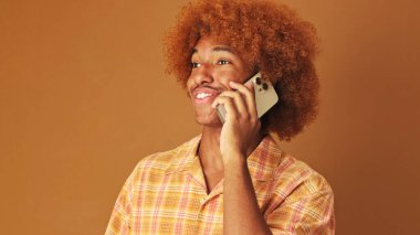 Gülümseyen stil sahibi kıvırcık çocuk cep telefonuyla konuşuyor. Stüdyoda kahverengi arka planda izole edilmiş.