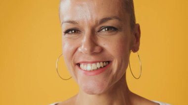 Kılsız genç bir kadının yakın plan portresi. Stüdyoda sarı arka planda izole edilmiş kameraya bakıp gülüyor.