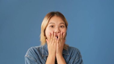 Kot gömlek giymiş, elleri stüdyoda mavi arka planda izole bir şekilde ağzını kapatan şaşırmış genç bir kadının yakın çekimi.