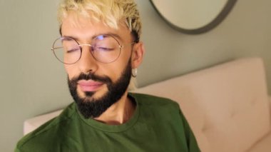 Yakın plan, sakallı genç sarışın adam rahat bir oturma odasında koltukta oturup kameraya bakıyor.