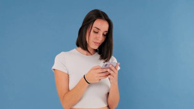 Beyaz elbiseli güzel esmer kadın cep telefonu parşömenleri kullanıyor. Stüdyoda mavi arka planda izole edilmiş online alışveriş yapıyor.