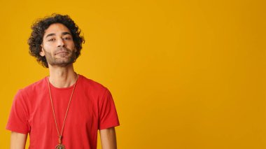 Kıvırcık saçlı, kırmızı tişörtlü çekici bir adam. Stüdyoda sarı arka planda izole edilmiş kameraya bakıyor.