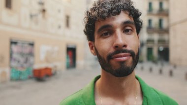 Yakından, sakallı, yeşil tişörtlü, sevimli esmer adam kameraya bakıyor.