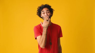 Kırmızı tişörtlü, kıvırcık saçlı mutlu çekici adam stüdyoda sarı arka planda izole edilmiş kameraya hava öpücüğü gönderiyor.