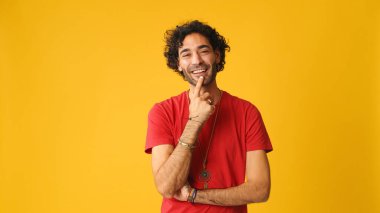 Kıvırcık saçlı, kırmızı tişörtlü, bir şeyler düşünen, sarı arka planda, stüdyoda izole edilmiş, dalgın, çekici bir adam.