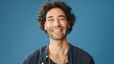 Yakın plan, kıvırcık saçlı, mavi gömlekli adam başını kaldırıyor ve stüdyonun mavi arka planında izole edilmiş kameraya bakıyor.