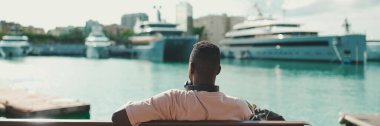 Genç Afrikalı adam limandaki yat ve gemi arka planında rahat oturuyor. Arka plan, Panorama