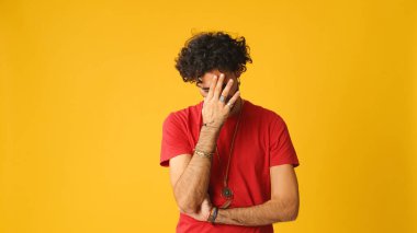 Dalgın, kıvırcık saçlı, kırmızı tişörtlü, çenesini ovuşturan ve stüdyoda sarı arka planda yaşayan çekici bir adam.