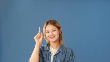 Kot gömlek giyen genç kadın, stüdyoda mavi arka planda izole edilmiş bir şekilde parmağını yukarı kaldırıyor.