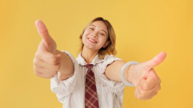 Gömlek ve kravat giymiş gülümseyen genç bir kadın stüdyoda sarı arka planda izole edilmiş kameraya çift parmak işareti yapıyor.