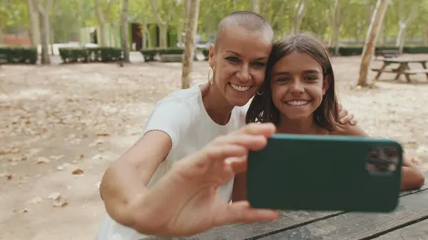 Happy mother and daughter sitting in the park taking selfie on mobile phone