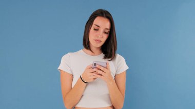 Beyaz elbiseli güzel esmer kadın stüdyoda mavi arka planda izole edilmiş cep telefonu kullanıyor.