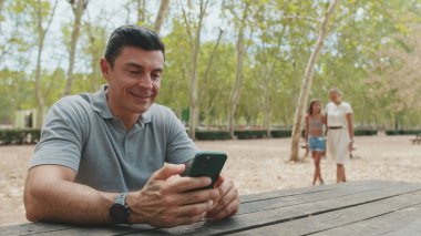 Orta yaşlı, gri tişörtlü bir adam parkta otururken elinde telefonla karısını ve kızını selamlıyor.