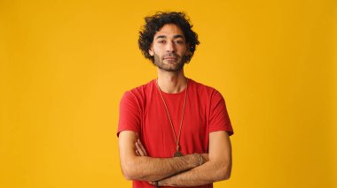 Kıvırcık saçlı, kırmızı tişörtlü, kollarını kavuşturup stüdyoda sarı arka planda izole edilmiş kameraya bakan çekici bir adam.