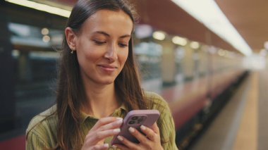 Kahverengi saçlı, zeytin yeşili süveter giyen genç bir kadın, tren istasyonunda dururken cep telefonu kullanıyor.